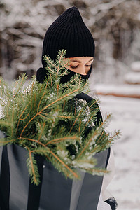 冬季风格摄影照片_新年前夜，一个穿着白色西装和巴拉克拉法帽的女孩在冬季森林里拿着一包圣诞树。新年的概念