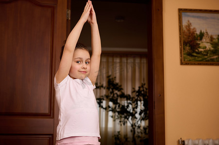 健身的小女孩摄影照片_漂亮的小女孩举起双臂，伸展身体，练习瑜伽，在室内锻炼时热身
