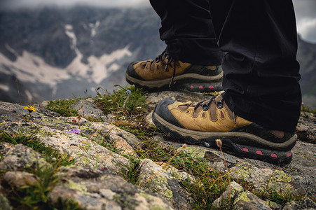 特写镜头，在雪山的背景下，穿着徒步靴的腿站在悬崖附近的一块石头上。