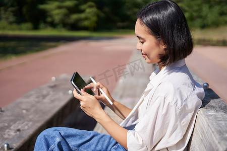 亚洲女孩，在公园里画画，在阳光明媚的日子里坐在街上，拿着数字图形平板电脑和笔，开心地笑着