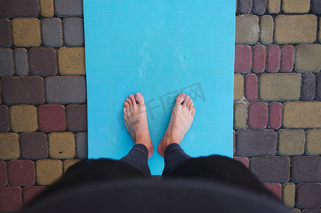 运动健身垫摄影照片_在活跃男子的脚正上方，瑜伽运动员赤脚站在蓝色健身垫上，同时在户外练习瑜伽