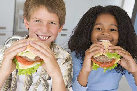 男孩吃汉堡摄影照片_两个小孩在厨房里微笑着吃芝士汉堡
