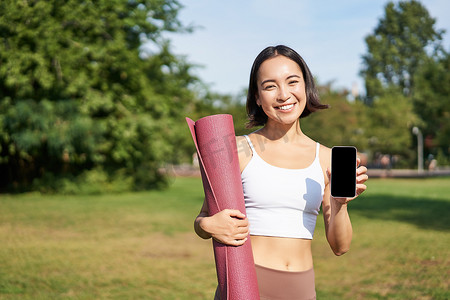 兴奋的健身女孩推荐运动和锻炼申请，展示手机屏幕，训练结束后站在公园里用橡胶瑜伽垫