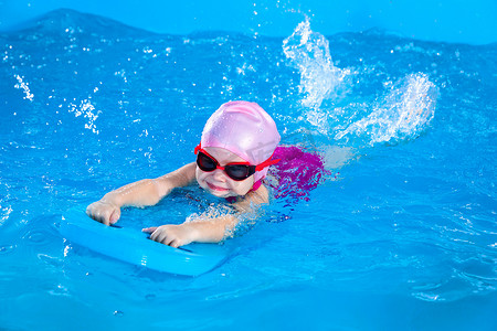室内游泳池摄影照片_学龄前可爱的女孩在室内游泳池里用颤动板学习游泳