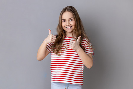 斜条纹底纹摄影照片_穿着条纹 T 恤的小女孩带着露齿的微笑看着镜头，竖起大拇指。