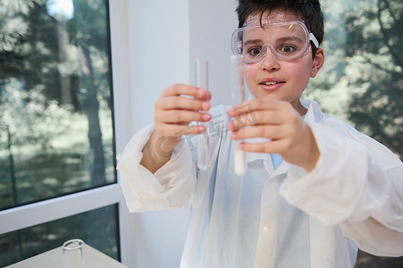 戴着护目镜的聪明男学生，穿着实验室外套，看着试管，着迷于正在进行的化学反应