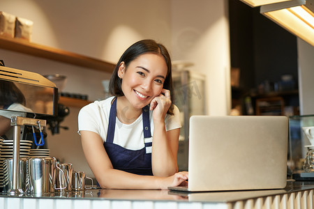 微笑的韩国女性的画像，咖啡店的咖啡师，拿着笔记本电脑站在柜台前，在她自己的咖啡店里微笑着，看起来自信、个体经营的女企业家