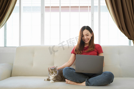 快乐的亚洲女人在家里和猫一起在舒适的沙发上使用笔记本电脑