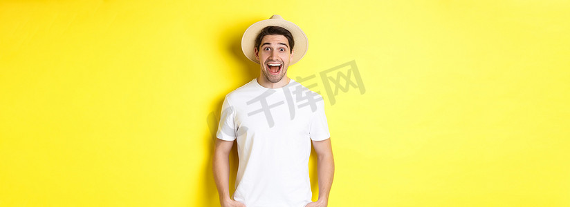 戴草帽的男摄影照片_惊讶的戴草帽的男游客看起来很开心，对旅行社的广告反应惊讶，站在黄色背景上