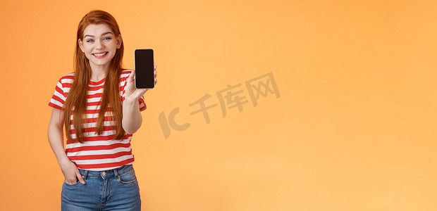 开朗帅气的 20 多岁红发女性介绍社交媒体特色产品手持智能手机、展示相机手机显示屏、推广设备应用、广泛微笑、橙色背景