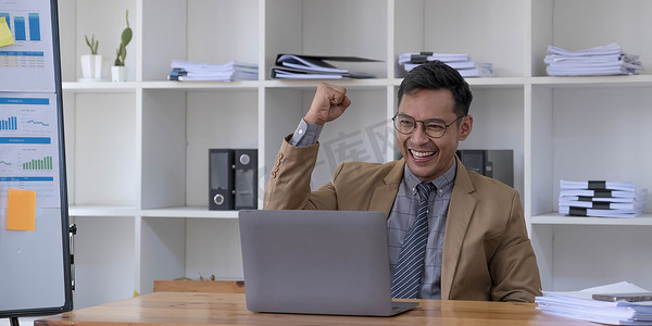 快乐的年轻商人穿着西装看着笔记本电脑，网上的好消息让他兴奋不已，幸运的成功赢家站在办公桌前，举起手来庆祝商业成功