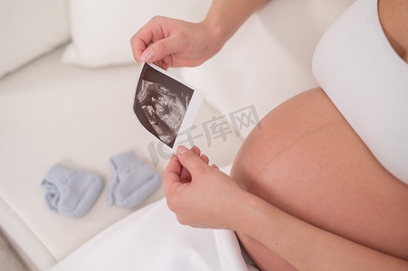 一位没有露面的孕妇坐在白色沙发上，手里拿着一张胎儿的超声波照片。