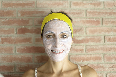 放松的女人在脸上涂矿物粘土面膜以恢复活力，头上有头带