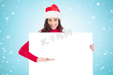 戴着圣诞老人帽子、微笑着的黑发女郎指向白色海报的合成图像