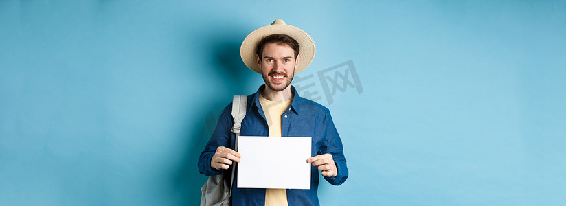 背包展示摄影照片_快乐的游客背着背包，展示着一张空纸，对着镜头微笑，站在蓝色背景上。