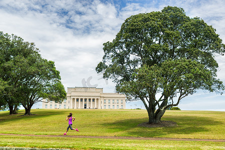 奥克兰市公园在奥克兰 Domain 公园与纪念博物馆的绿草和树木上跑步的慢跑女孩。