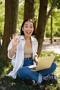 亚洲年轻女性的画像，学生做作业，在公园工作，带着笔记本电脑坐在树旁，展示好的标志，批准 smth