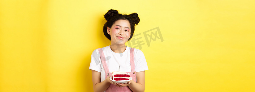 生日快乐的亚洲女孩，妆容鲜艳，在蛋糕上吹蜡烛，许愿，站在黄色背景上