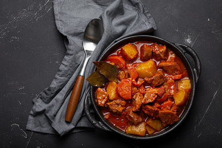 深色直纹摄影照片_牛肉炖土豆、胡萝卜和美味的肉汁放在黑色砂锅锅里，用月桂叶和勺子放在黑色深色乡村混凝土背景上，从上面看。