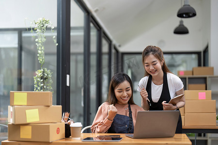 快乐的两位亚洲女企业家，在家庭办公室用智能手机查看在线购物商店的订单后，为销售成功而微笑，在线商业和电子商务的概念