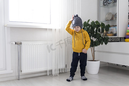 一个身穿黄色毛衣、头戴灰色帽子的小孩站在带恒温器的加热器附近。