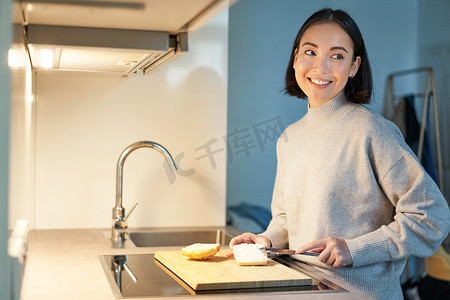 可爱的亚洲女人自己烤面包，切面包，在厨房里准备三明治