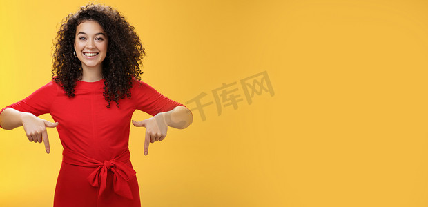 黄墙摄影照片_工作室拍摄了一张令人愉快的微笑的年轻卷发女性，她穿着红色的裙子，用食指向下指着，作为宣传令人敬畏的活动，广泛地微笑着建议尝试或购买黄墙