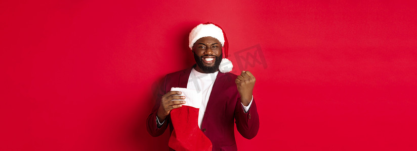 快乐的男人欢欣鼓舞，在圣诞袜子里收到礼物，握拳，满意地微笑，站在红色背景上