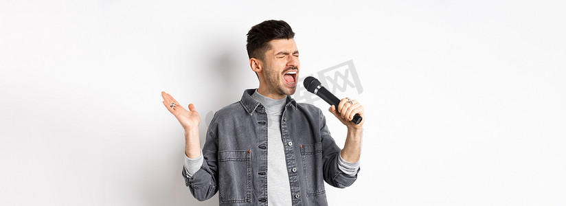 男人唱歌摄影照片_帅哥用麦克风热情地唱歌，为观众表演，拿着麦克风和手势，站在白色背景上
