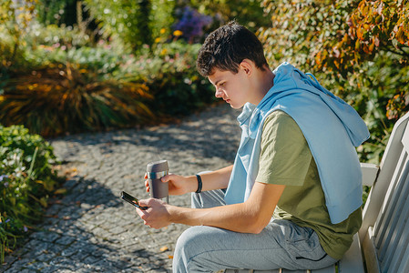 一个少年坐在秋天公园的长椅上，喝着保温杯里的咖啡，看着手机。