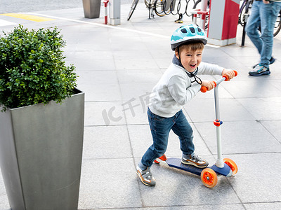 生态停车场摄影照片_戴着头盔的蹒跚学步的人在停车场骑着滑板车，停着自行车。