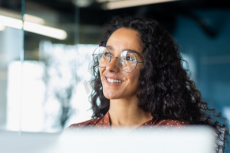 办公室里戴着眼镜的成功女商人的肖像，微笑着看着窗外的女人，梦想着对成就结果感到满意的女人，西班牙裔女人在工作场所带着笔记本电脑