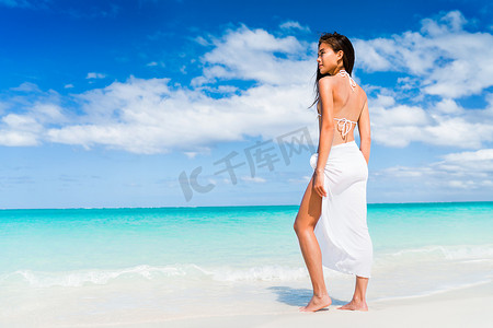 穿着白色沙滩装比基尼和时尚围裙的优雅亚洲女性站在海滩上。