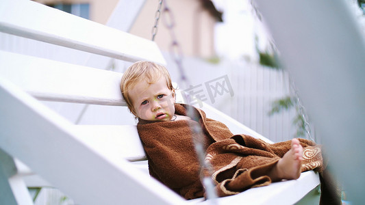 悲伤的贫穷小女孩摄影照片_一个一岁大的小女孩，裹着毛巾，脸脏兮兮的，在夏天孤独地坐在花园里的秋千上。