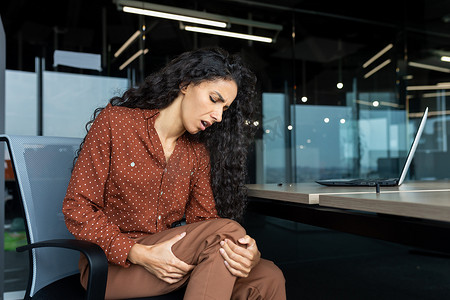 在办公室工作的拉丁美洲女性，女商人有严重的腿痛，坐在椅子上的桌子上按摩肌肉，在工作中使用笔记本电脑