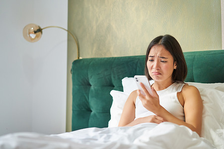 忧心忡忡的亚洲女性拿着手机，接到不好的电话，看起来忧心忡忡，躺在床上电话交谈困难