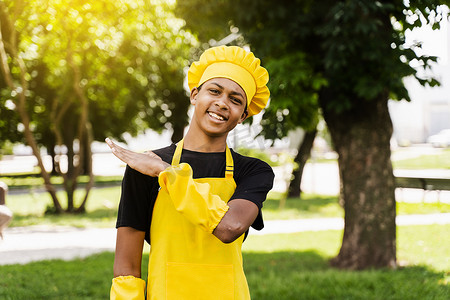 身着厨师帽和黄色围裙制服的快乐黑人非洲少年厨师在户外微笑。