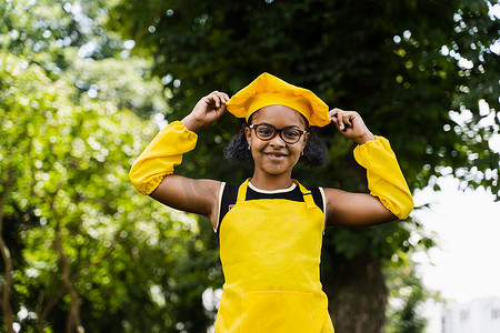 黑人厨师摄影照片_身穿厨师帽和黄色围裙制服的非洲黑人儿童厨师女孩摸着她的厨师帽。