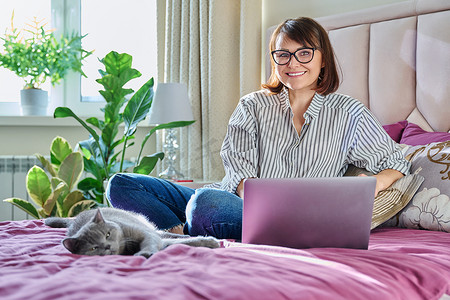 沙发猫摄影照片_家里的中年妇女躺在床上，带着笔记本电脑和猫