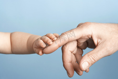 婴儿蓝色背景摄影照片_蓝色背景下婴儿抱着男人手指的特写