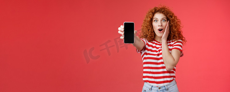 逗乐的红发帅气的卷发兴奋女孩分享很棒的社交媒体页面显示智能手机屏幕惊奇触摸脸颊激动影响很棒的应用程序红色背景