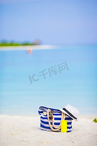 防晒沙滩摄影照片_白色沙滩上的蓝色包、草帽、太阳镜和防晒瓶