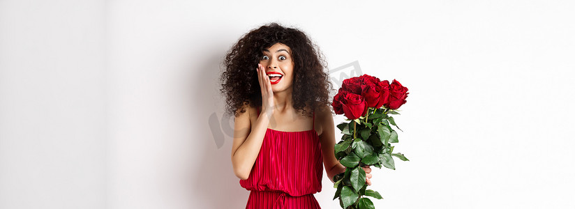 身着红色裙子、兴奋的卷发女人，收到一束玫瑰，看起来很惊讶，为浪漫的礼物而欢欣鼓舞，站在白色背景上