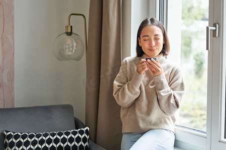 美丽的亚洲年轻女性坐在窗边喝咖啡，拿着浓咖啡杯，面带轻松、微笑的表情看着外面