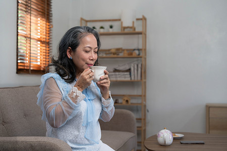 肖像，放松而平静的 60 多岁亚洲女性喝着热茶，在她的客厅里喝下午茶。