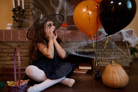 小女巫，看着气球，坐在南瓜和带糖果的篮子旁边，靠着带蜘蛛网的壁炉