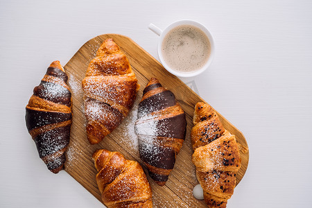 白桌木板上放着一杯咖啡和一堆开胃的棕色和巧克力羊角面包，上面放着糖粉，平铺