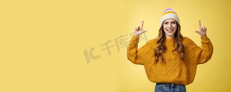 促销风摄影照片_友善迷人、精力充沛的 20 多岁年轻女性打扮时尚的帽子毛衣，在介绍促销时很有趣，指着举起的食指展示顶级广告，开心地笑着黄色背景
