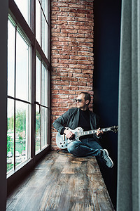 男歌手坐在窗台上，戴着耳机，吉他在家庭录音室录制曲目