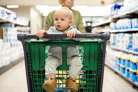 在超市杂货店的部门过道里，妈妈推着购物车带着她的婴儿男婴。
